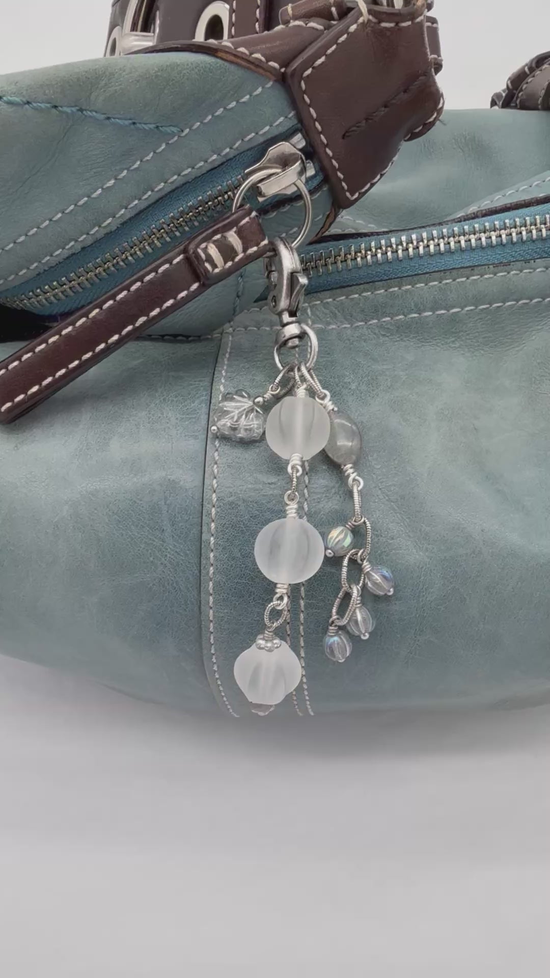 Video of Snowfall Serenade handmade bag charm with three frosted white fluted Lucite beads, a labradorite oval, gray Czech glass beads, a clear glass maple leaf, and an antique-silver-finish swivel clasp on a light blue leather handbag.