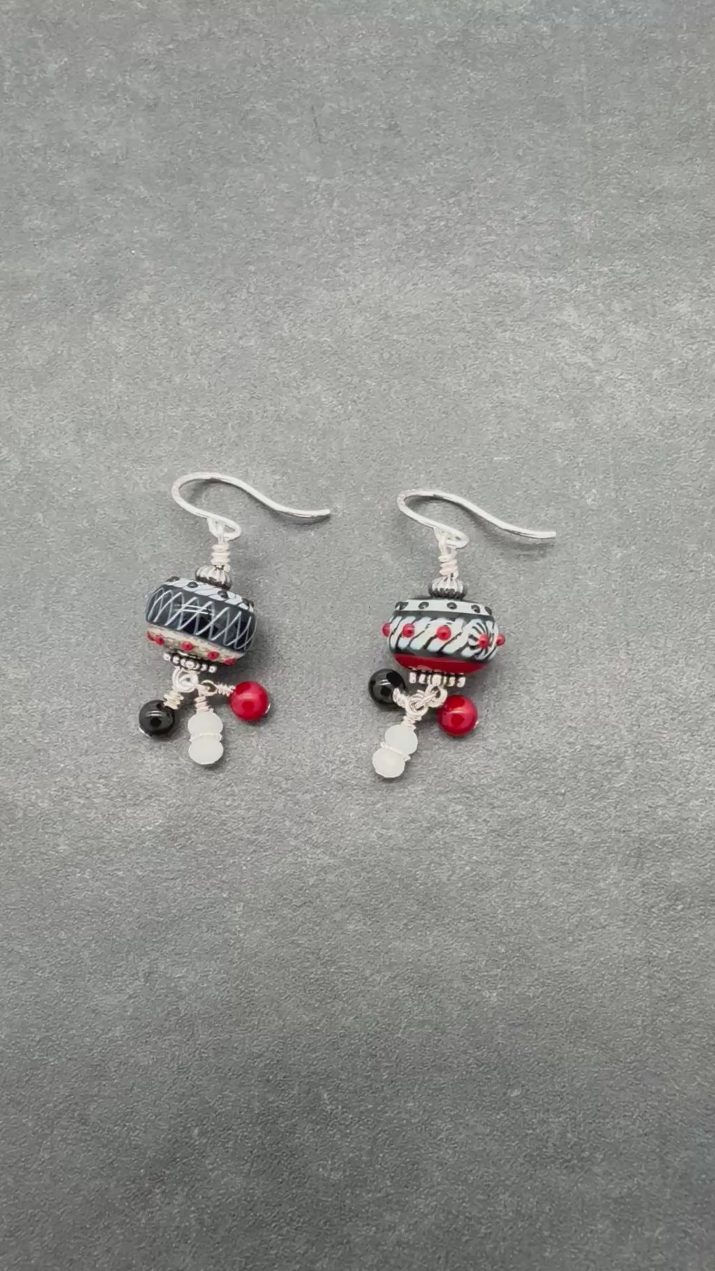 A video of a pair of asymmetrical earrings with silver ear wires and black and white patterned lampwork beads on a gray surface - made with love in Colorado by Ruby Acorn.