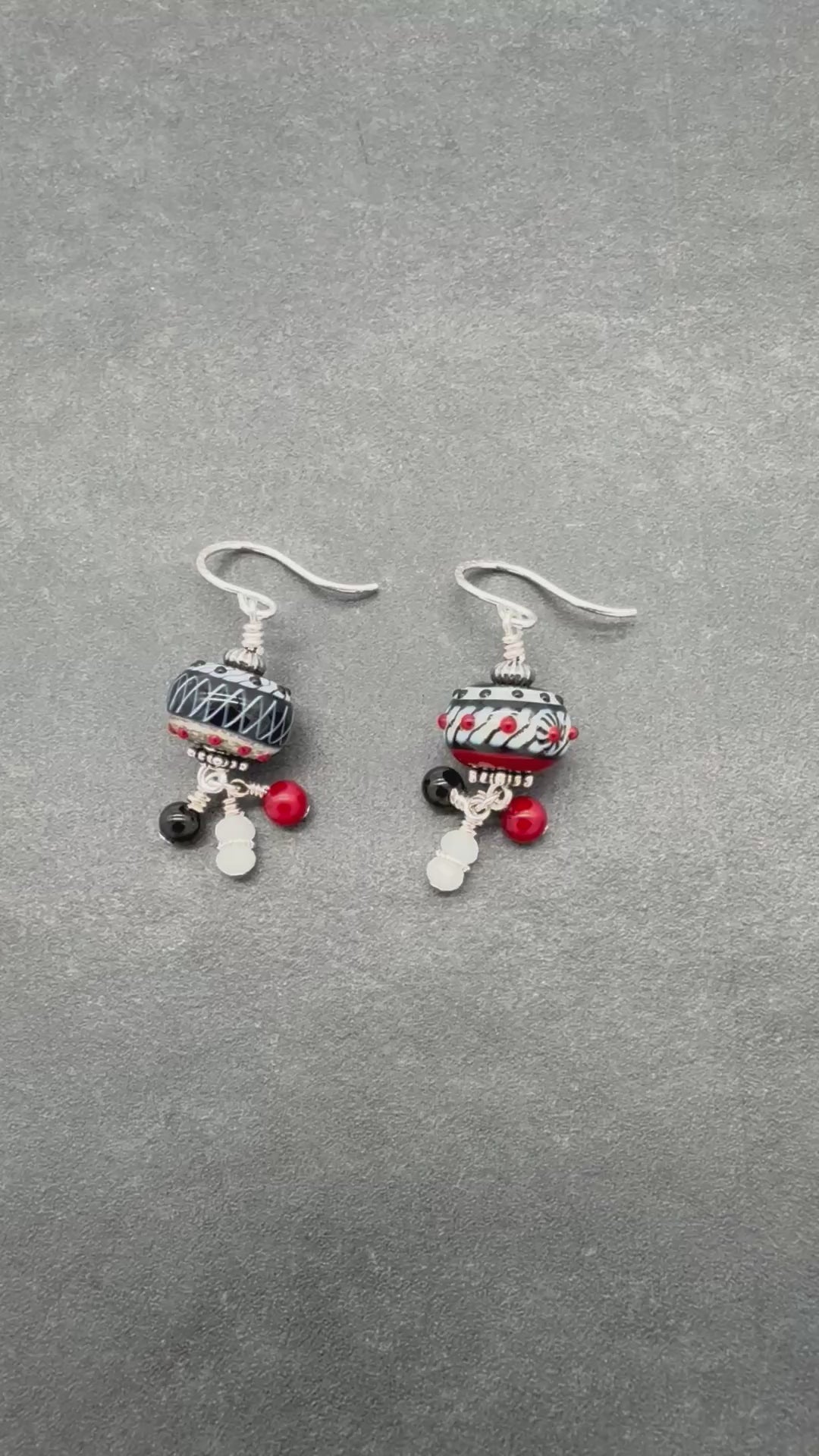 A video of a pair of asymmetrical earrings with silver ear wires and black and white patterned lampwork beads on a gray surface - made with love in Colorado by Ruby Acorn.