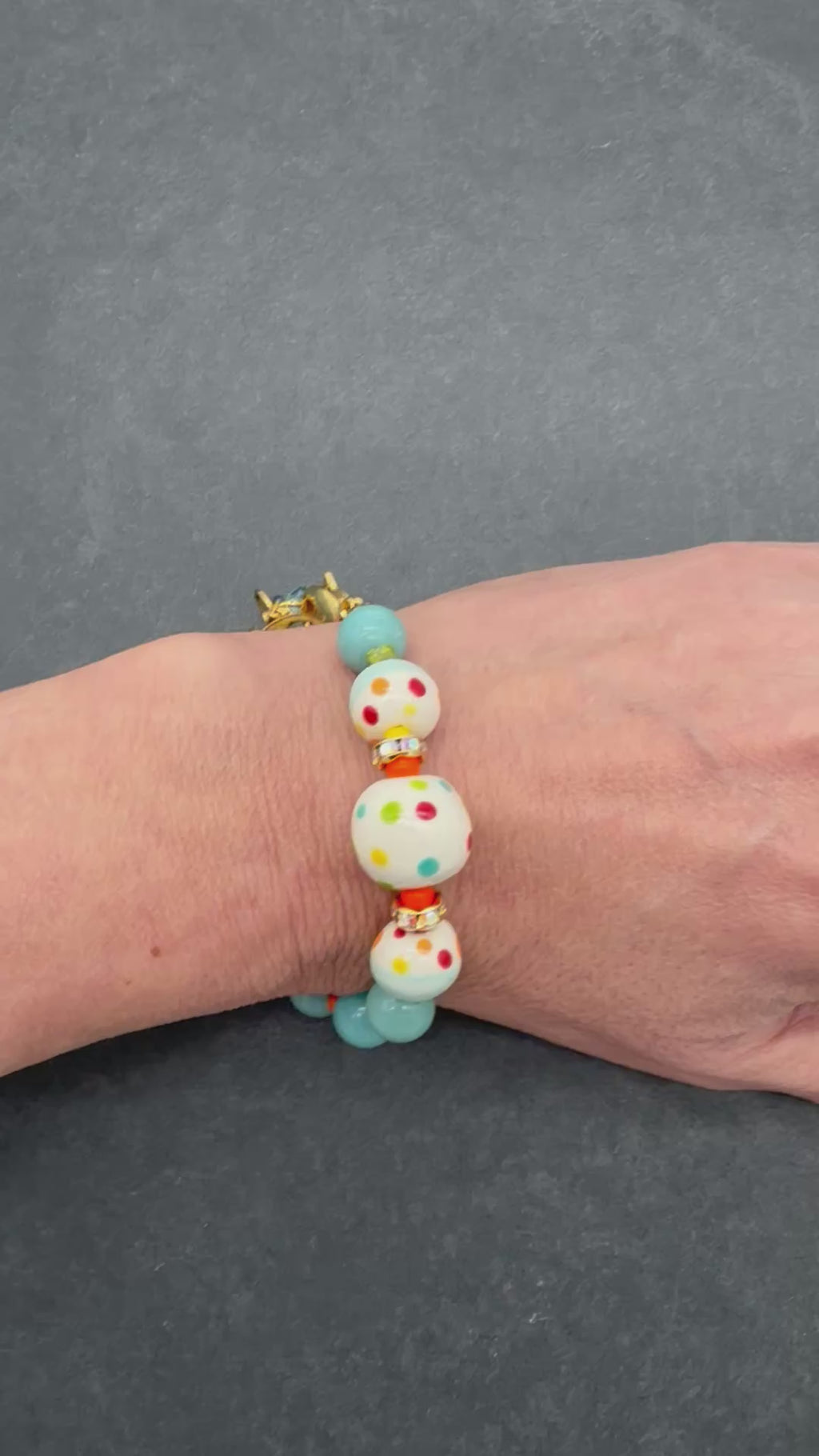 A video of a close-up of a person's hand wearing a beaded bracelet. The bracelet features light blue beads, white beads with colorful polka dots, and small orange spacer beads.