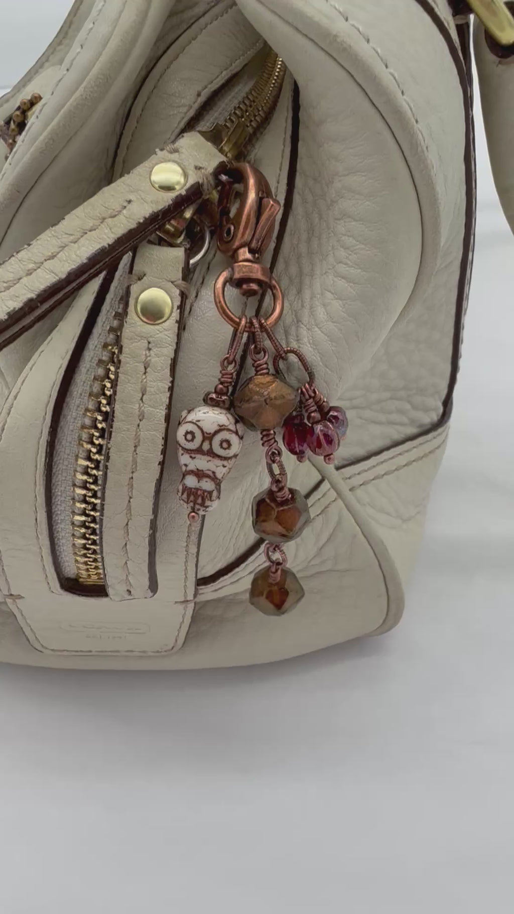 A video close-up of a cream handbag with a decorative copper-toned keychain charm featuring faceted brown beads, burgundy fluted melons, and a small owl - made with love in Colorado by Ruby Acorn.