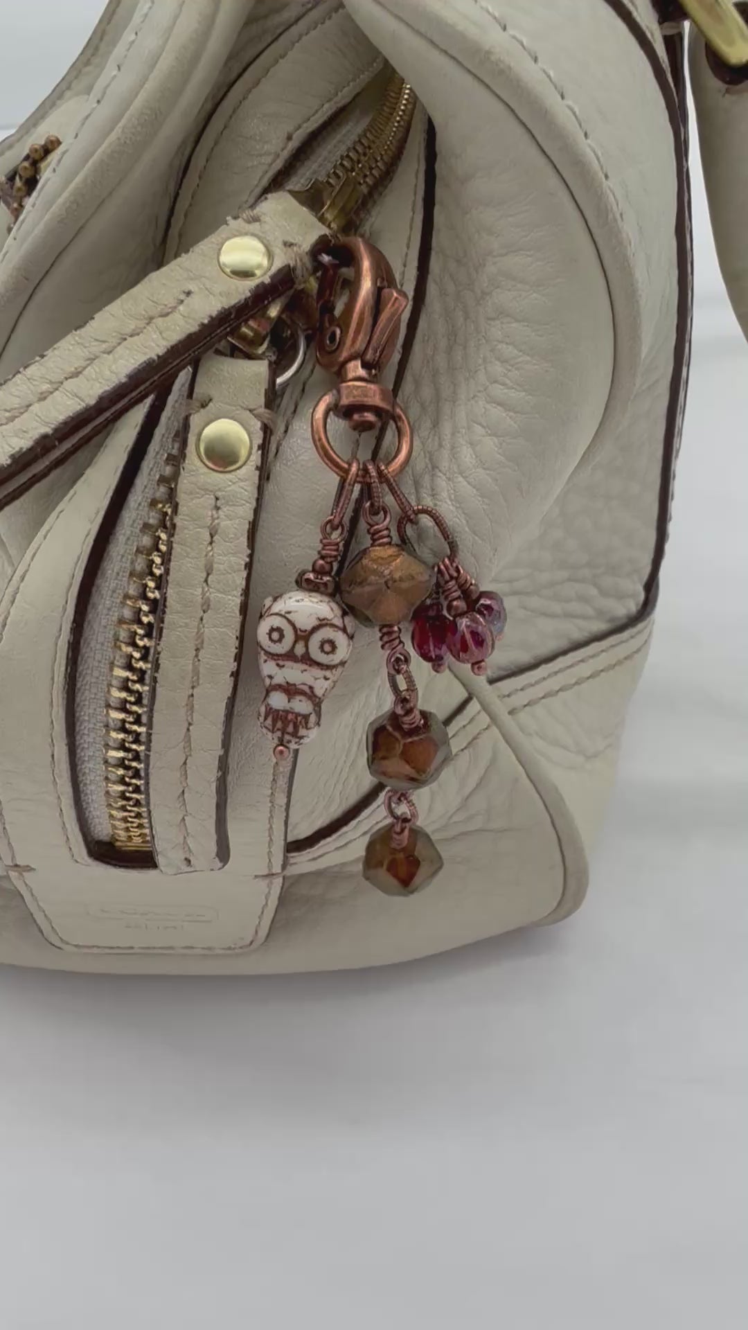 A video close-up of a cream handbag with a decorative copper-toned keychain charm featuring faceted brown beads, burgundy fluted melons, and a small owl - made with love in Colorado by Ruby Acorn.