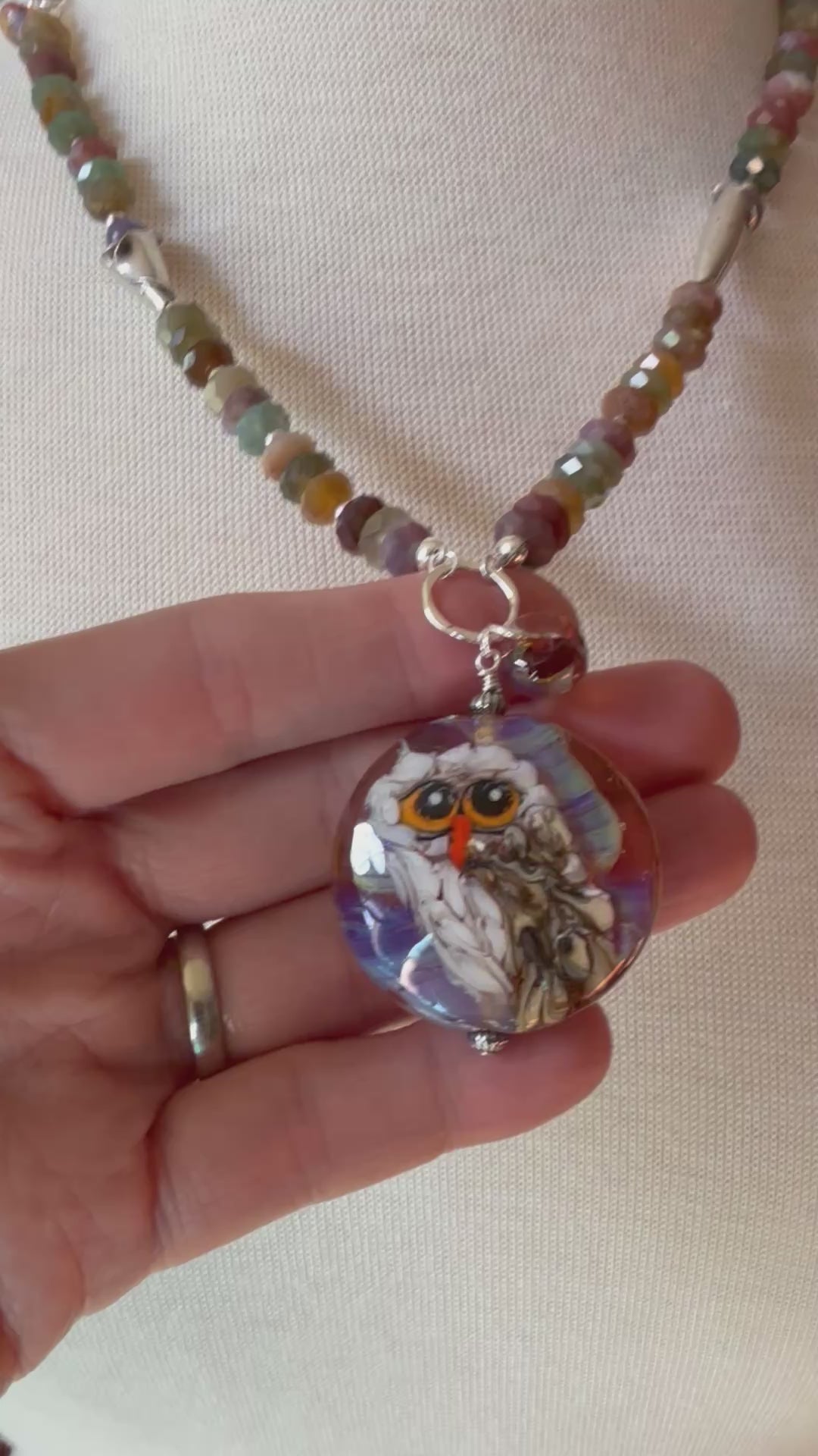 Video of a beaded necklace displayed on a cream fabric mannequin bust. The necklace features a strand of small, multicolored beads in earthy tones, and an artisan lampwork glass pendant depicting an owl with orange eyes and a purple, white, and brown patterned background - made with love in Colorado by Ruby Acorn.