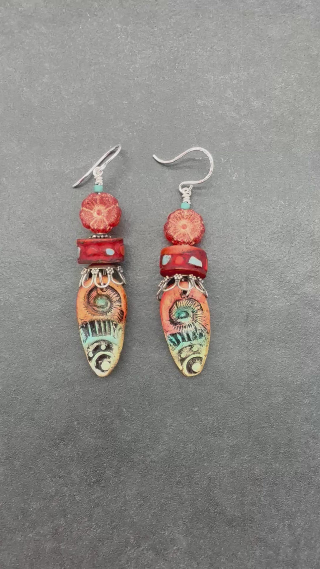 A Video of a pair of handmade dangle earrings featuring textured red and orange beads, turquoise accents, and silver-tone hooks on a gray background - made with love in Colorado by Ruby Acorn.