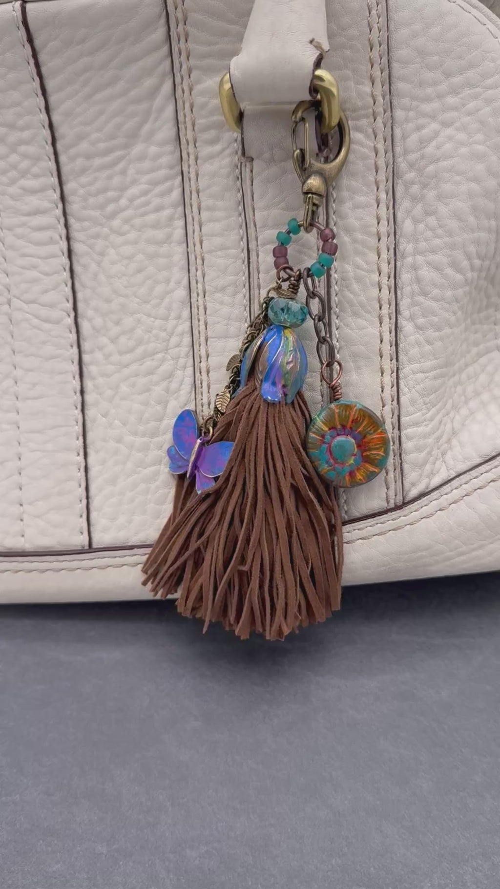 Video of decorative keychain with leather tassel and hand painted brass on a cream colored purse