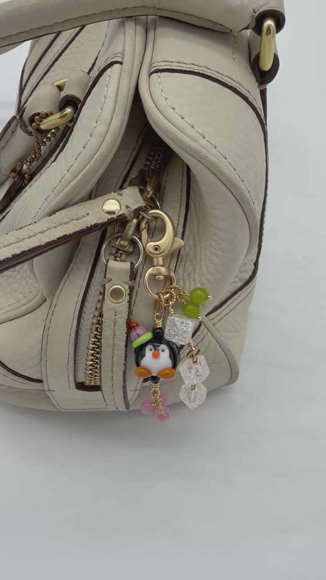A video of a close-up of a handbag zipper pull featuring a penguin charm with a green hat, clear faceted beads, and pink beads on a gold-tone clasp, attached to a light beige textured leather bag.