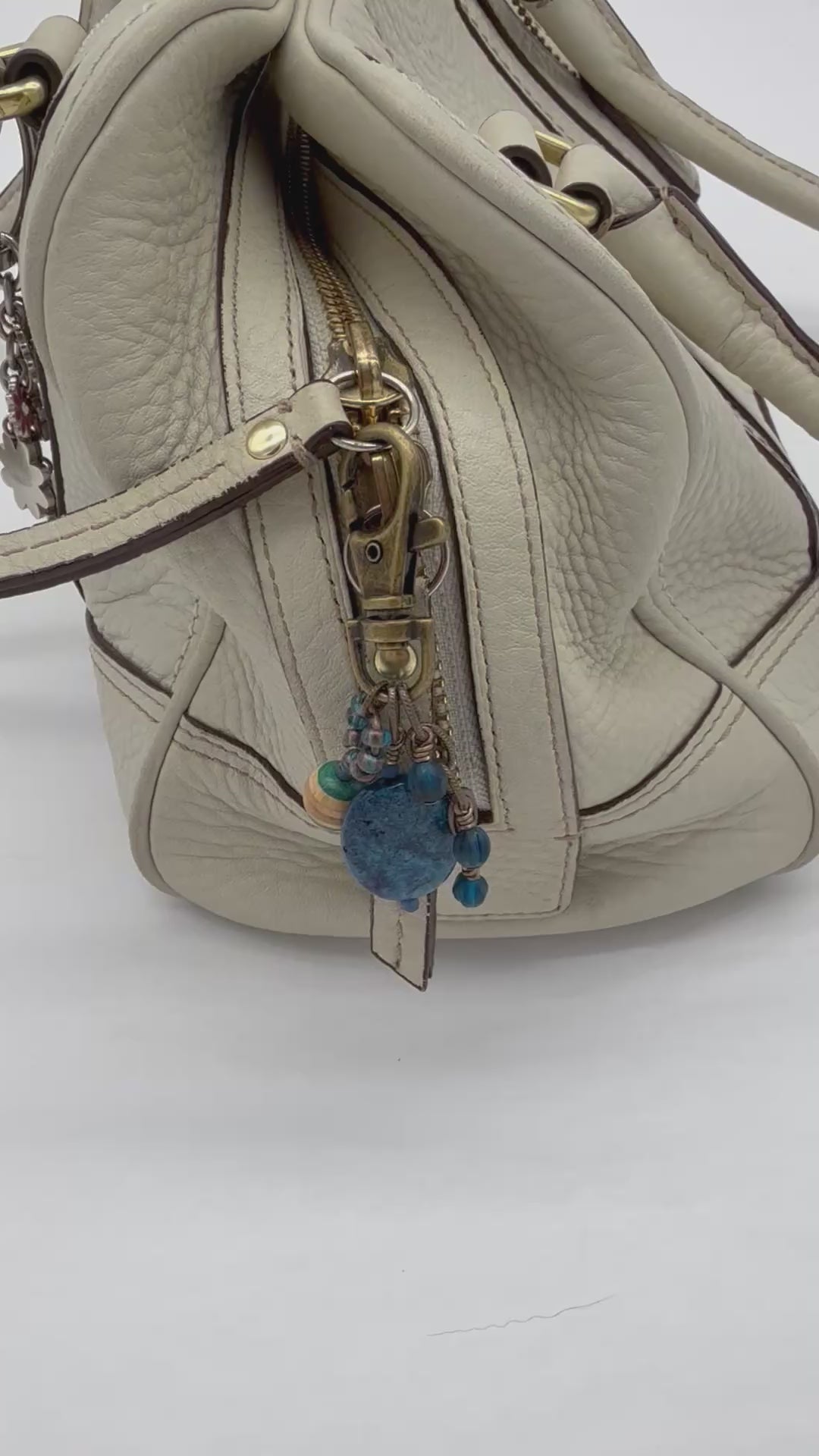 A video of a cream-colored textured leather handbag featuring a brass-tone lobster clasp and a decorative hanging charm with blue beads and wooden accents - made with love in Colorado by Ruby Acorn.
