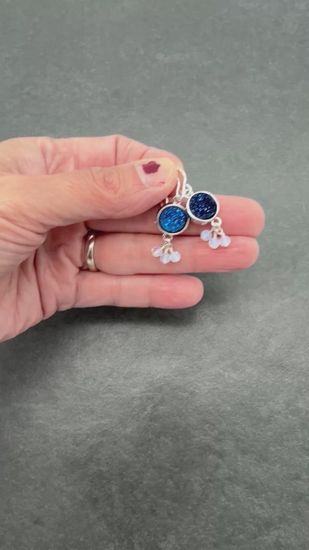 A video of a hand holding a pair of silver dangle earrings with round indigo crystal druzy links and small iridescent white beads.