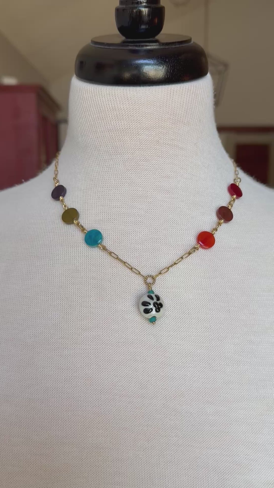 A video of a beaded necklace displayed on a white fabric mannequin bust. The necklace features a gold-tone chain with multiple colorful round beads in dark purple, olive green, light blue, red, and orange, ending in a small black-and-white paw print pendant - made with love in Colorado by Ruby Acorn.