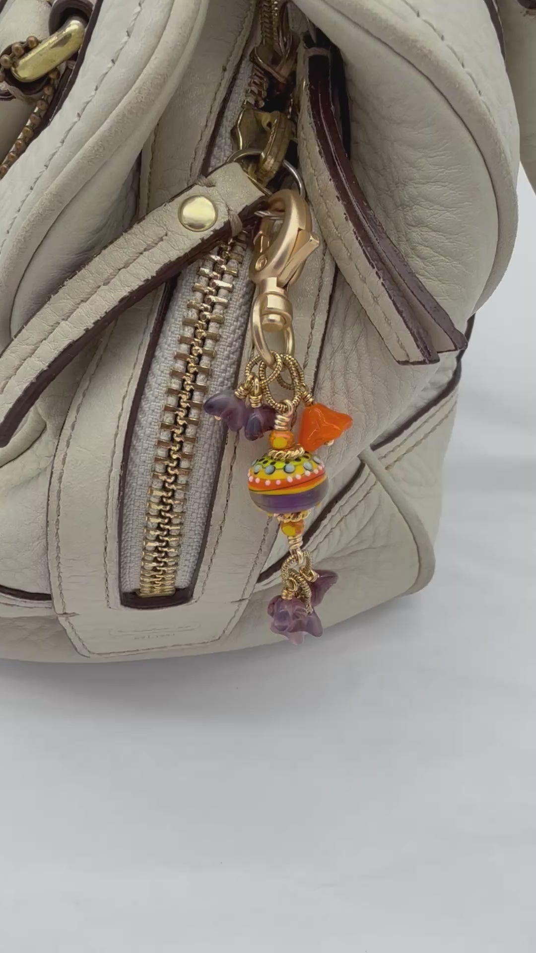 A video of a handbag charm with a gold lobster clasp, orange, yellow and purple lampwork bead, an orange bellflower, and purple flowers fastened on a cream purse - made with love in Colorado by Ruby Acorn.