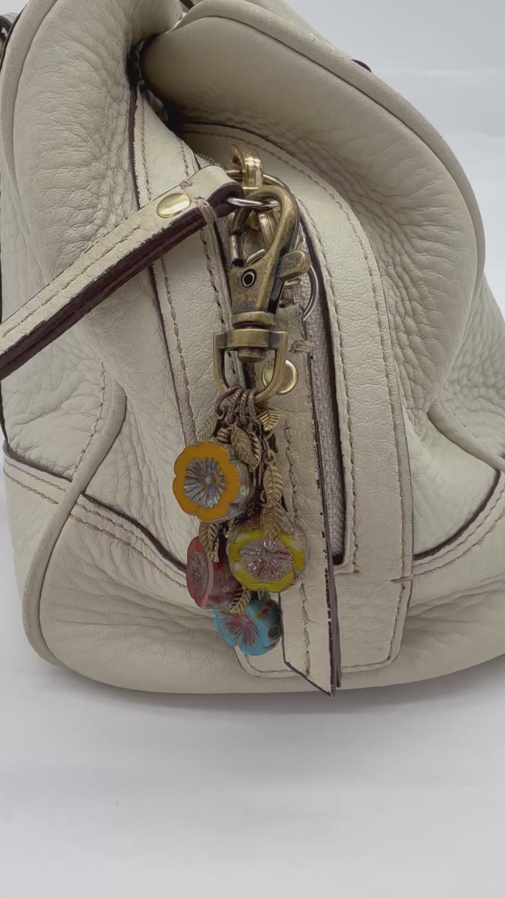A video close-up of a cream-colored leather handbag featuring a brass-tone zipper pull adorned with colorful floral charms in orange, red, blue, and green - made with love in Colorado by Ruby Acorn.