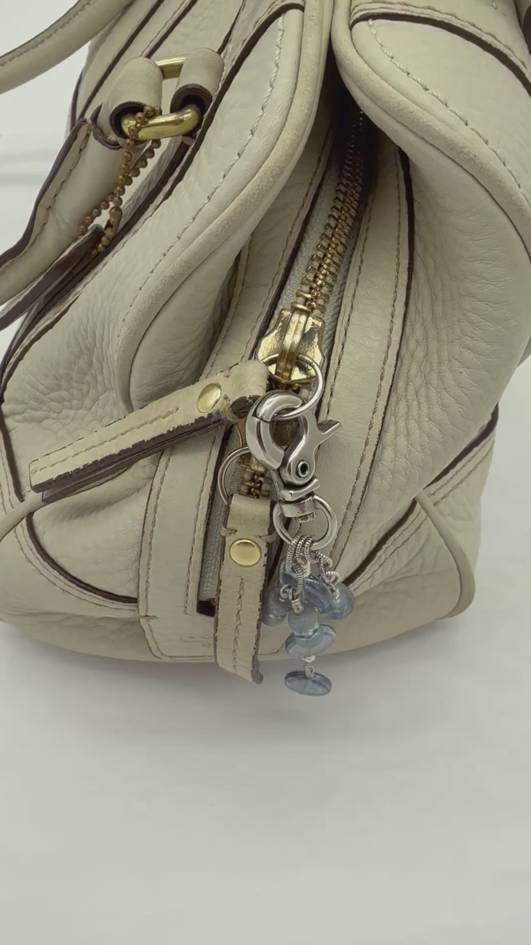 A video of a silver-toned beaded keychain or bag charm with round blue-gray beads and wire-wrapped accents, shown on a cream leather handbag.