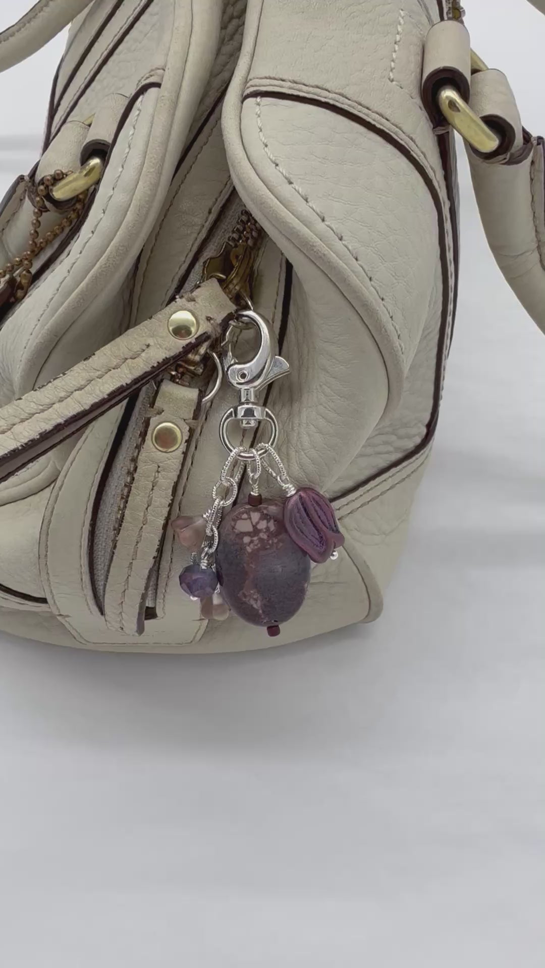 A video close-up of a handbag with a cream-colored leather zipper and strap, decorated with a silver-tone keychain charm featuring purple and dark brown marbled beads - made with love in Colorado by Ruby Acorn.
