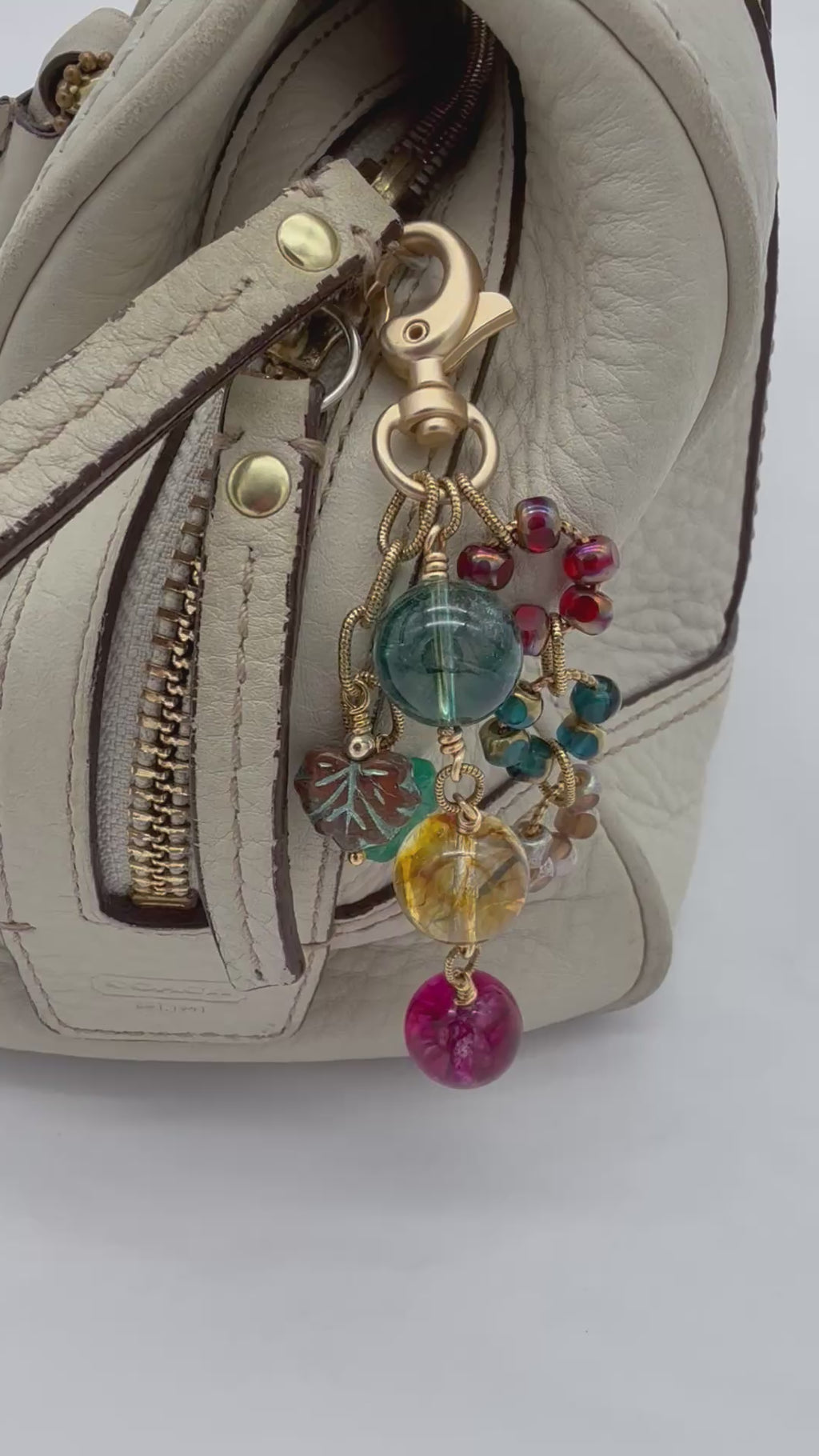 Video of a colorful handbag charm displayed on a cream leather handbag made with rainbow agate and Czech glass and a gold toned jumbo lobster clasp.
