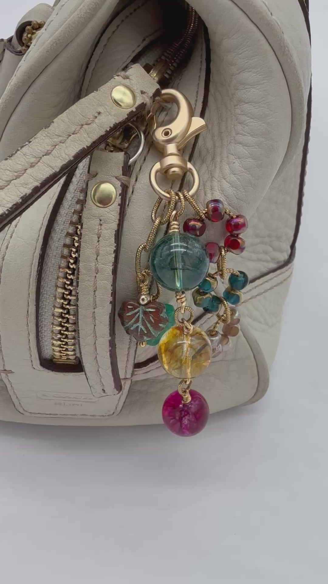 Video of a colorful handbag charm displayed on a cream leather handbag made with rainbow agate and Czech glass and a gold toned jumbo lobster clasp.