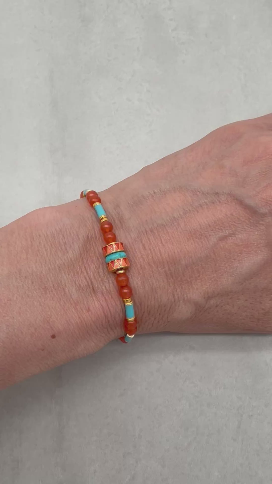 Video of a person's hand wearing a beaded bracelet featuring round coral-orange beads, light blue turquoise beads, and gold spacer rings, with a central decorative cylindrical bead and a gold clasp, shown on a neutral light gray background - made with love in Colorado by Ruby Acorn.