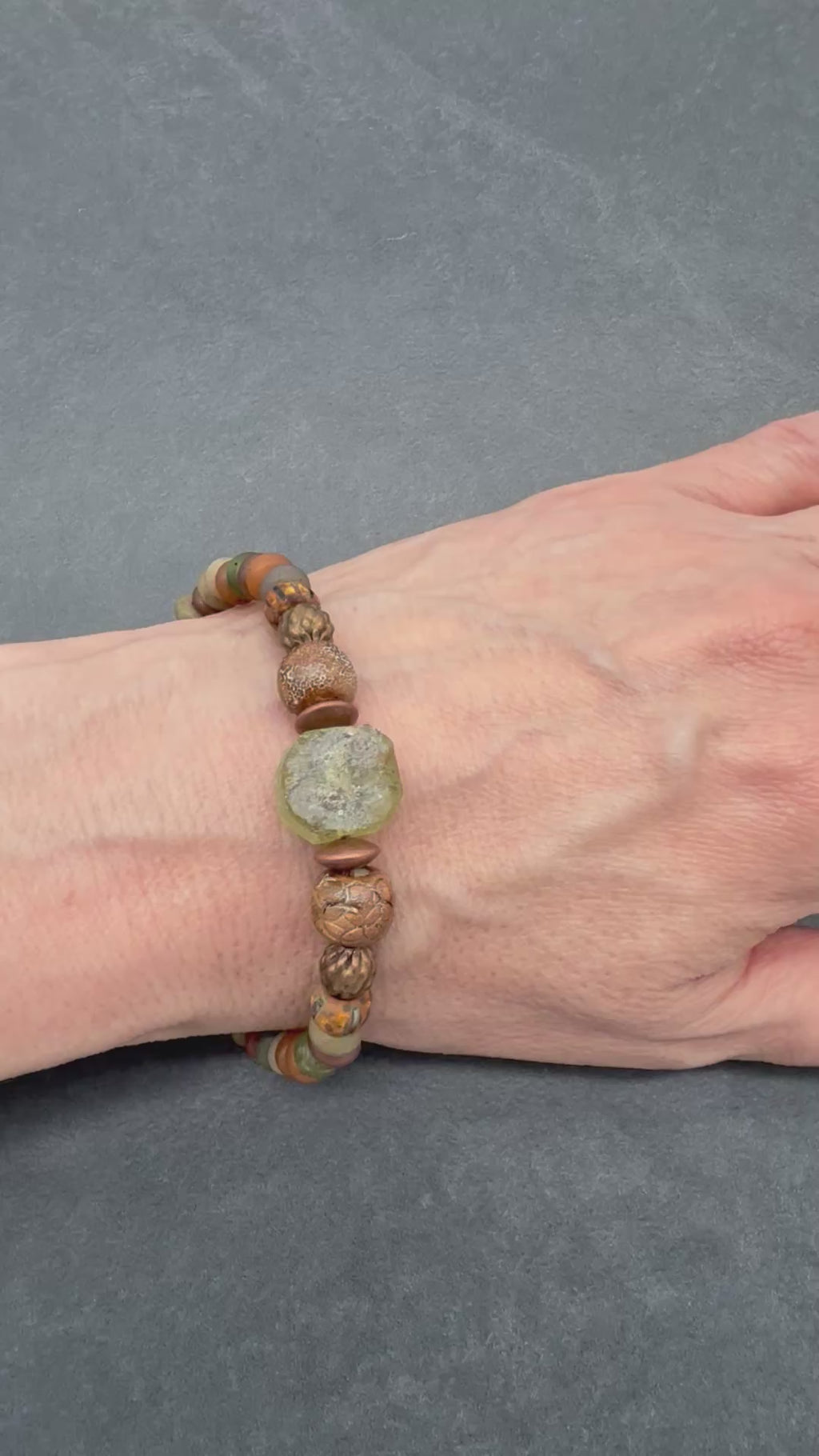 Video of a close-up of a hand resting on a dark gray surface, wearing a beaded bracelet made of brown and green beads - made with love in Colorado by Ruby Acorn. 