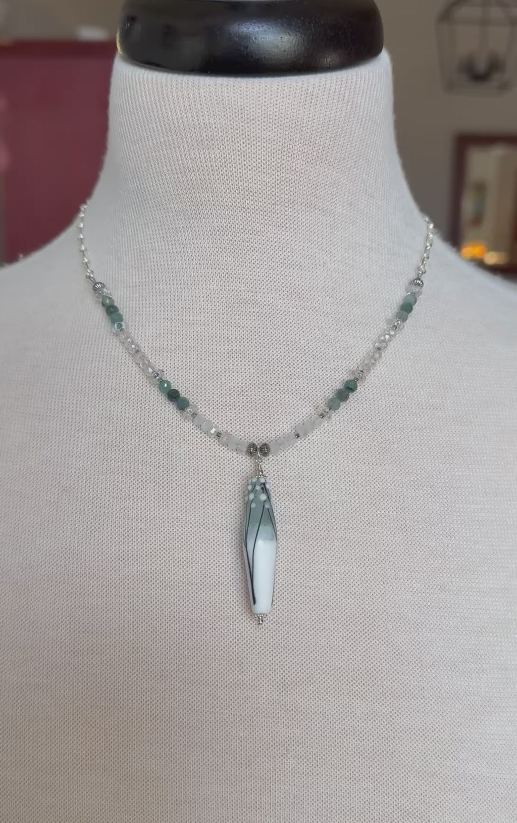 A video of a beaded necklace with a long tapered barrel artisan lampwork glass pendant displayed on a cream fabric mannequin bust. The necklace features a silver-tone chain with clear faceted beads, small dark green beads, and the focal pendant.