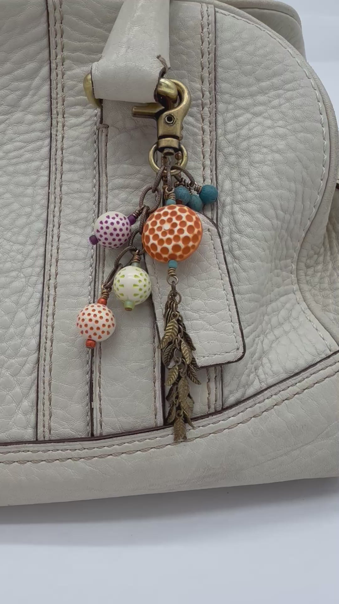 A video of a close-up of a cream-colored textured leather bag featuring a decorative keychain or bag charm attached near the zipper. The charm includes a brass-tone clasp, small blue beads, patterned vintage Lucite beads in orange, green, and purple, and a dangling bronze-toned leaf tassel - made with love in Colorado by Ruby Acorn.