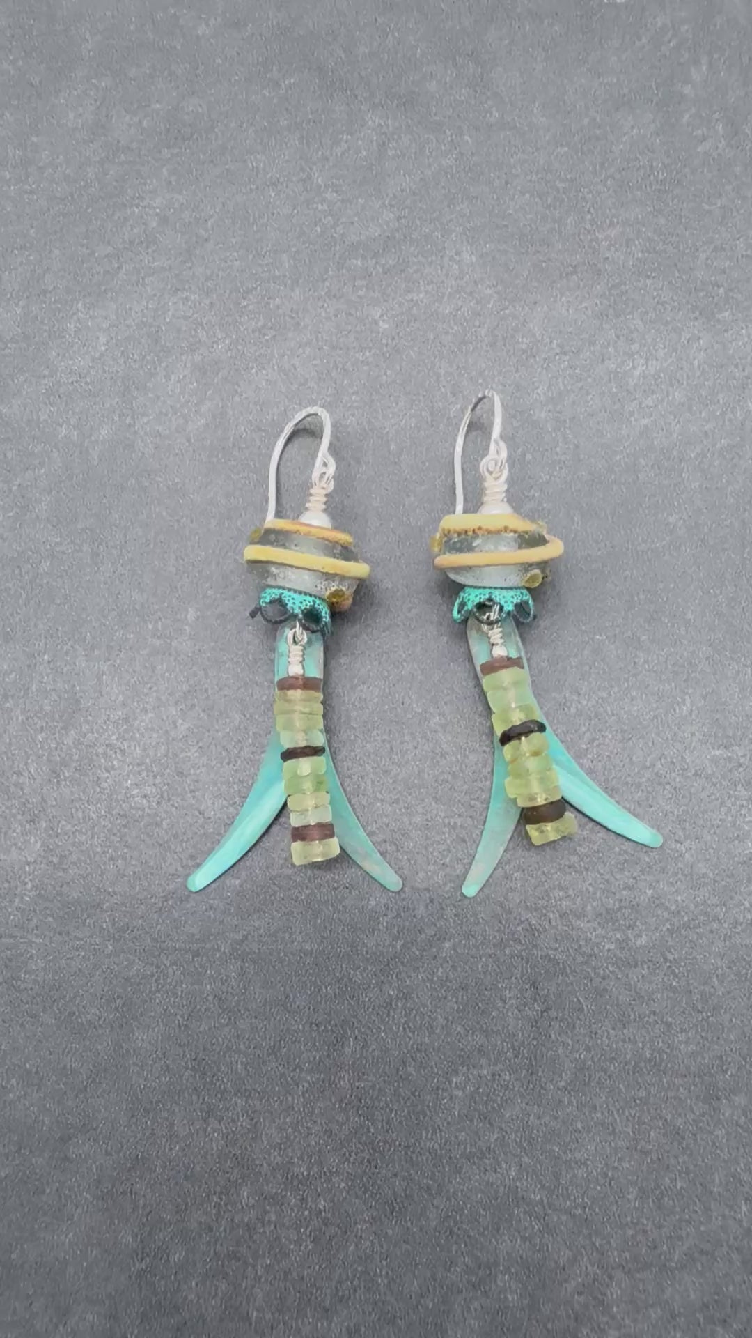 A video of a pair of handmade dangle earrings displayed on a gray background. The earrings feature silver ear wires, artisan lampwork glass, ancient Roman glass, and verdigris daggers - made with love in Colorado by Ruby Acorn.
