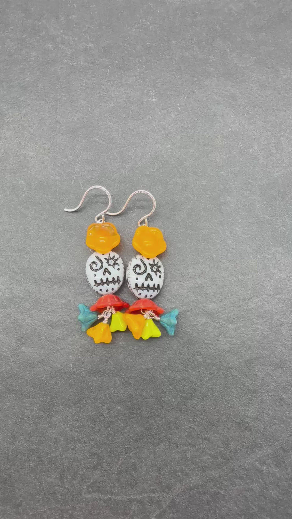 A video of a pair of handmade dangle earrings featuring silver-tone fishhook wires, translucent orange flower beads, white beads with black sugar skull designs, and colorful flower charms in red, yellow, green, and blue at the bottom, displayed on a dark gray textured background - made with love in Colorado by Ruby Acorn.