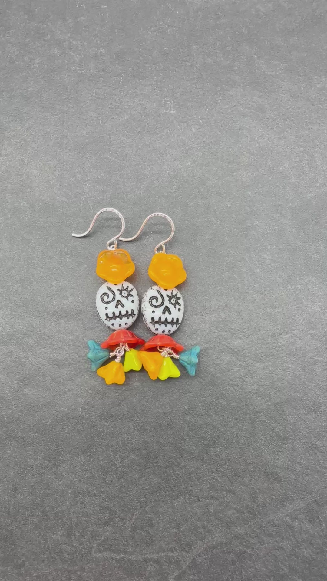 A video of a pair of handmade dangle earrings featuring silver-tone fishhook wires, translucent orange flower beads, white beads with black sugar skull designs, and colorful flower charms in red, yellow, green, and blue at the bottom, displayed on a dark gray textured background - made with love in Colorado by Ruby Acorn.