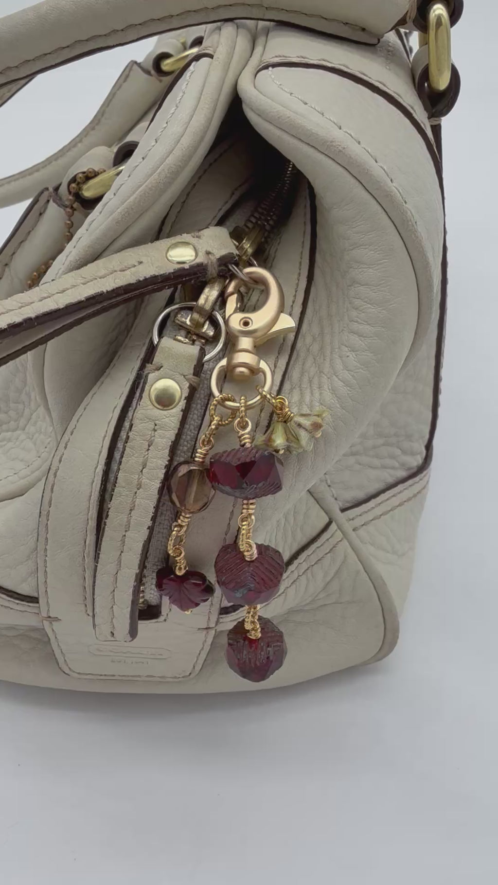 A video of a close-up of a cream-colored leather handbag featuring a decorative gold-tone keychain charm with faceted dark red beads and small floral accents.