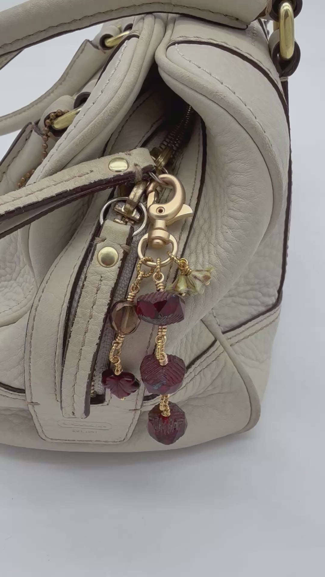 A video of a close-up of a cream-colored leather handbag featuring a decorative gold-tone keychain charm with faceted dark red beads and small floral accents.