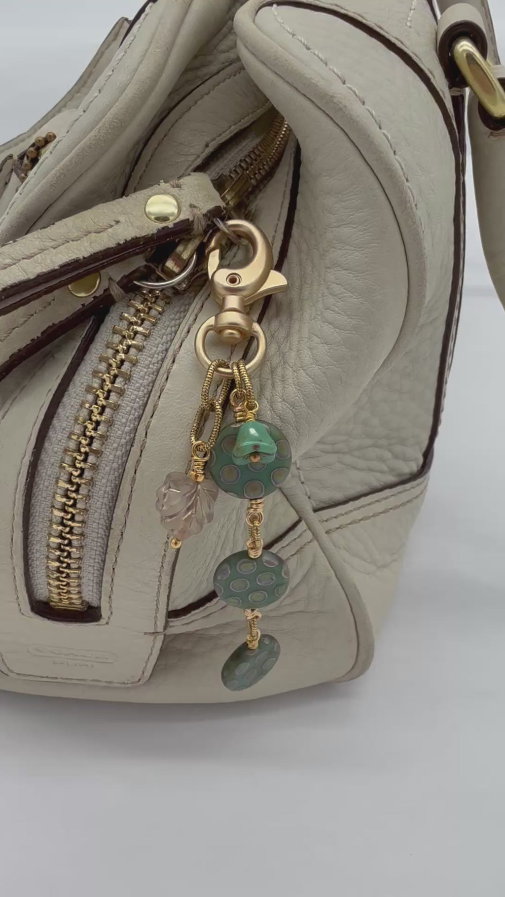 A video close-up of a handbag in light beige leather with a gold zipper, featuring a decorative keychain charm with green patterned discs, a flower bead, and a small iridescent leaf charm - made with love in Colorado by Ruby Acorn.
