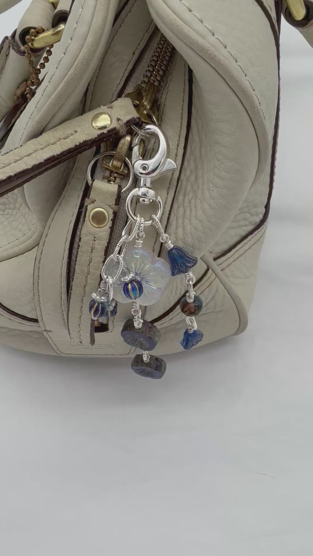 A video of a close-up of a handbag in light beige textured leather with a silver-tone keychain charm featuring a white iridescent flower and dark blue faceted beads.