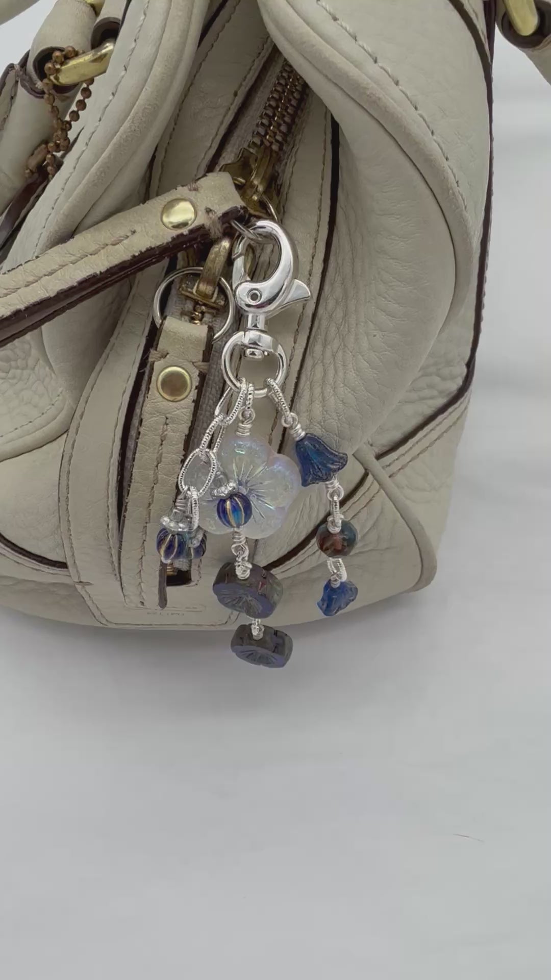 A video of a close-up of a handbag in light beige textured leather with a silver-tone keychain charm featuring a white iridescent flower and dark blue faceted beads.