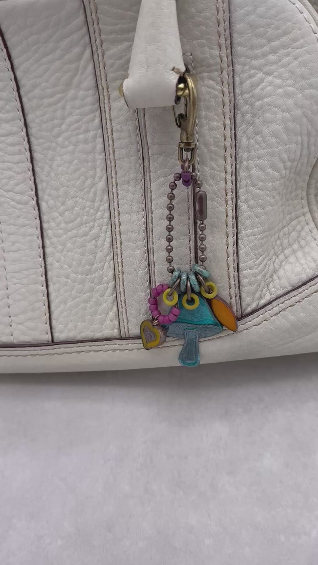 Video of a colorful keychain with a mushroom charm hanging on a cream colored leather handbag