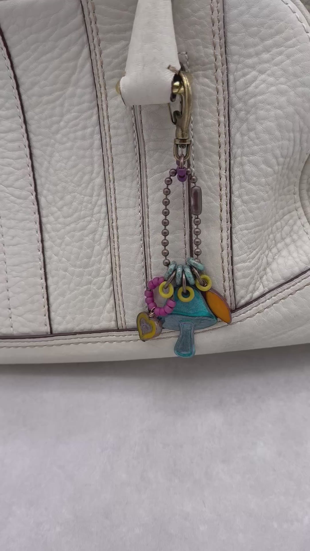 Video of a colorful keychain with a mushroom charm hanging on a cream colored leather handbag