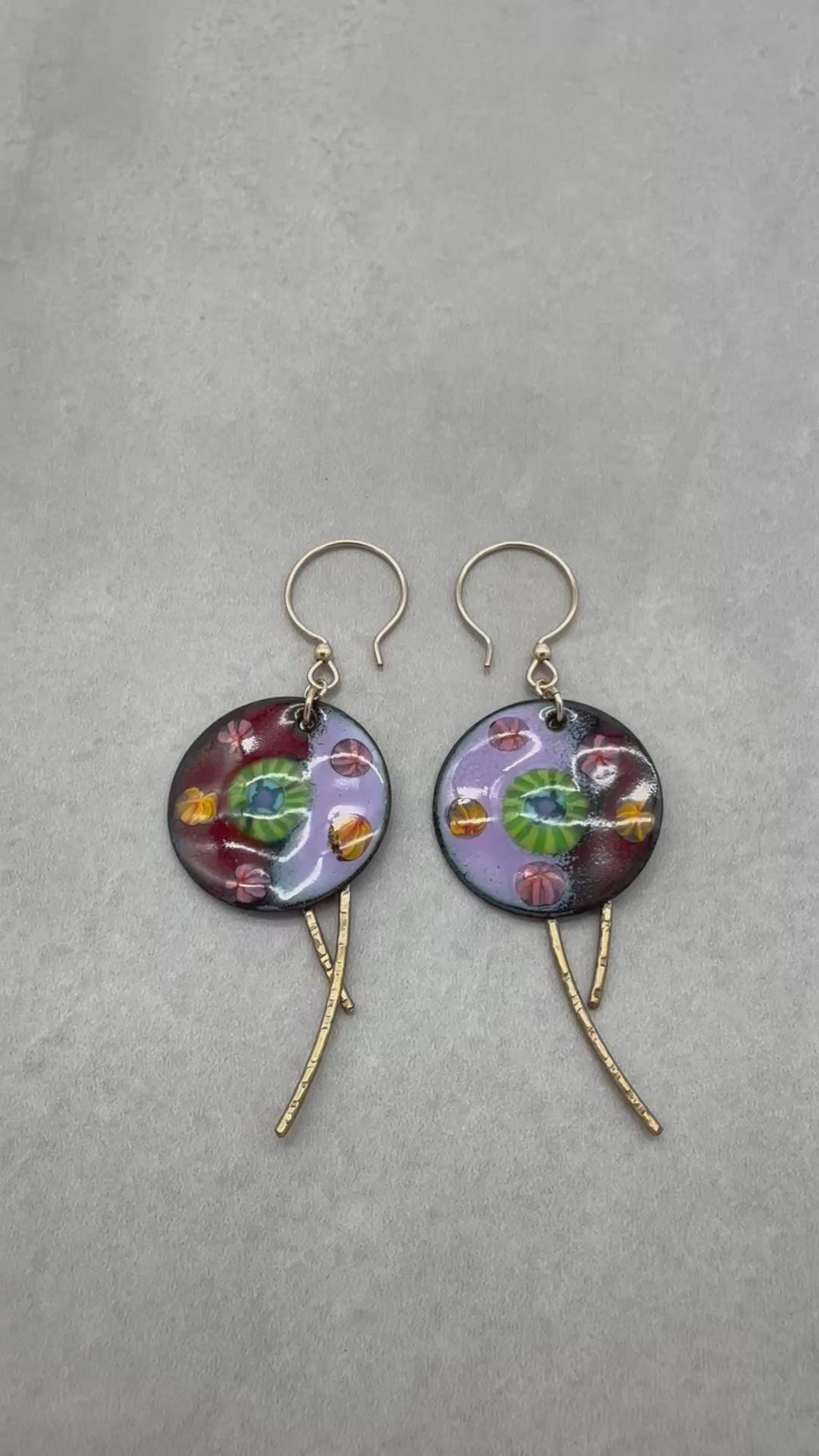 A video of a pair of handmade enamel earring pendants with gold-tone ear wires and brass textured daggers, featuring a colorful Millefiori pattern on a light background - made with love in Colorado by Ruby Acorn.