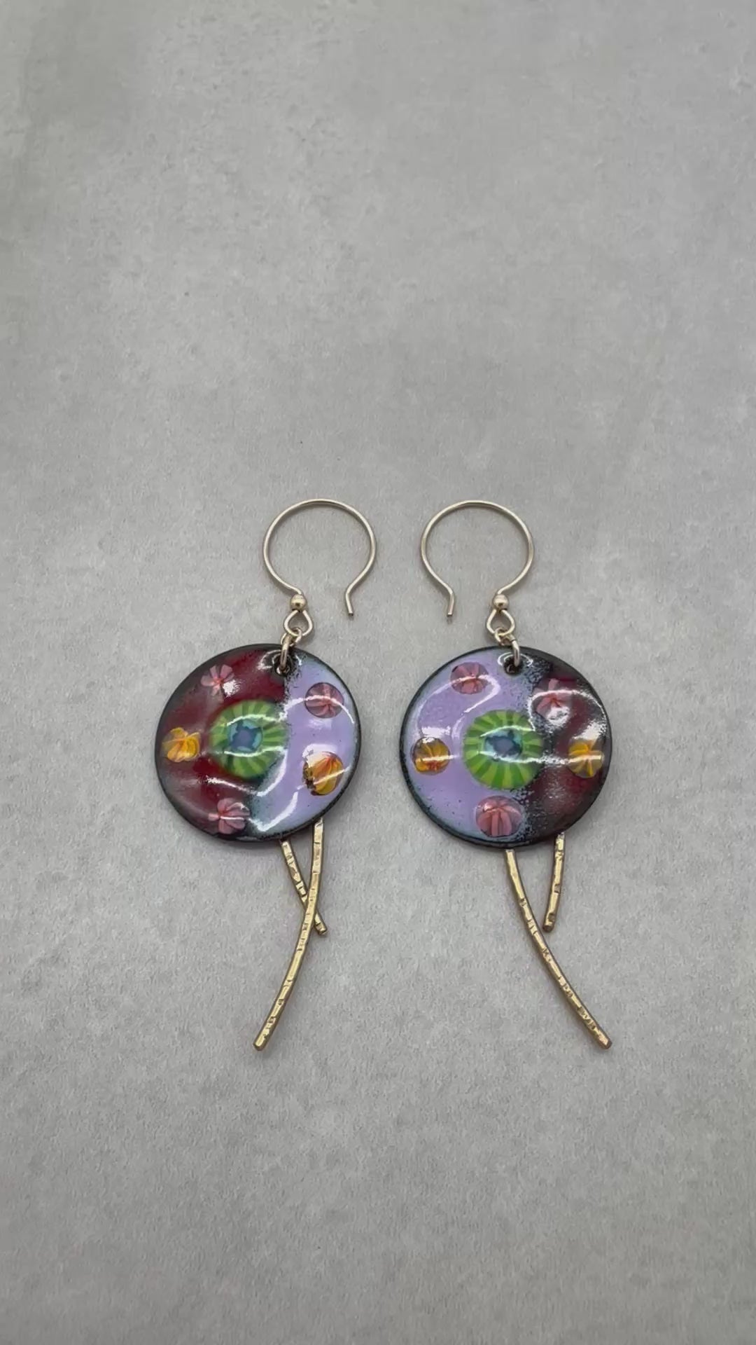 A video of a pair of handmade enamel earring pendants with gold-tone ear wires and brass textured daggers, featuring a colorful Millefiori pattern on a light background - made with love in Colorado by Ruby Acorn.