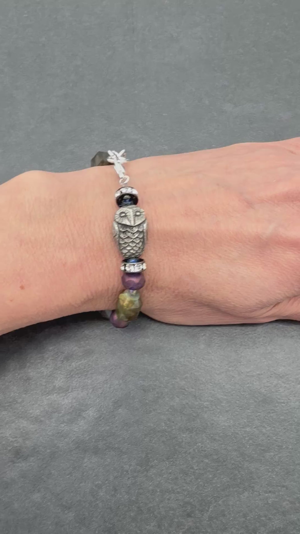 A video of a silver-toned beaded bracelet with an owl charm and purple beads, shown on a person's wrist against a dark gray textured background.