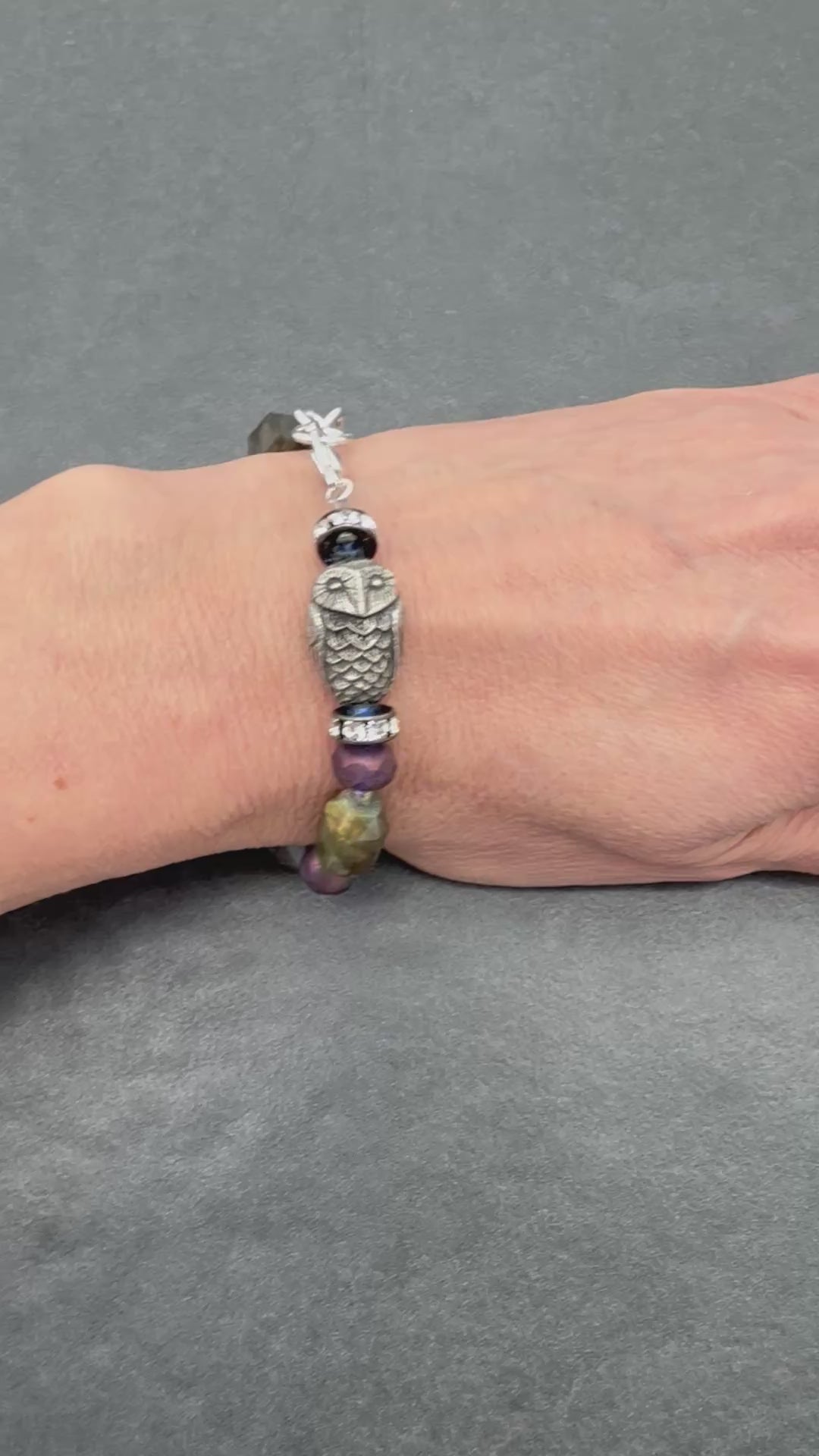 A video of a silver-toned beaded bracelet with an owl charm and purple beads, shown on a person's wrist against a dark gray textured background.