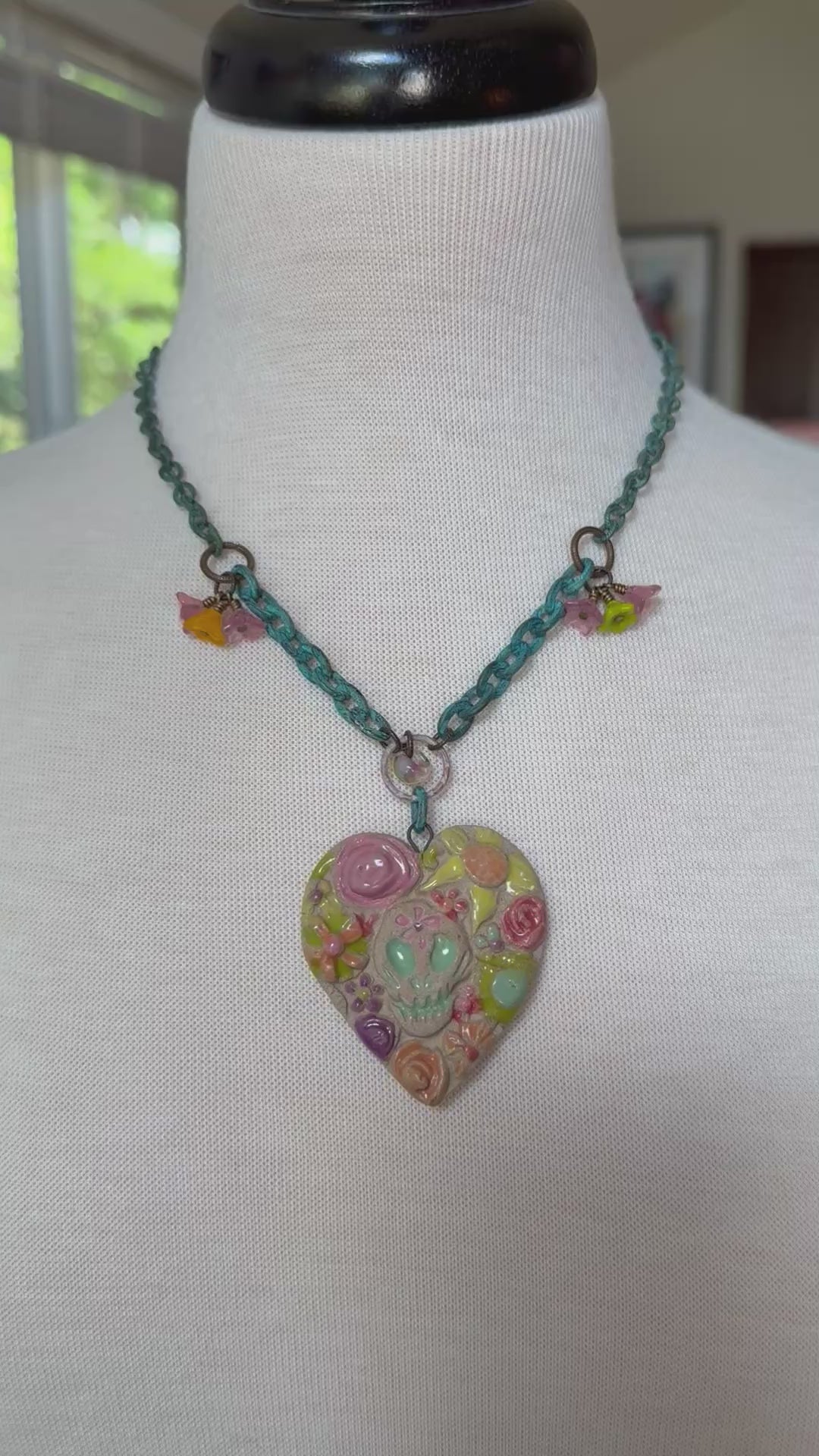 Video of a colorful heart-shaped Dia de los Muertos pendant necklace displayed on a white fabric mannequin bust. The necklace features a verdigris chain with small dangling bellflower charms and a large heart pendant decorated with a pastel sugar skull and floral patterns - made with love in Colorado by Ruby Acorn.
