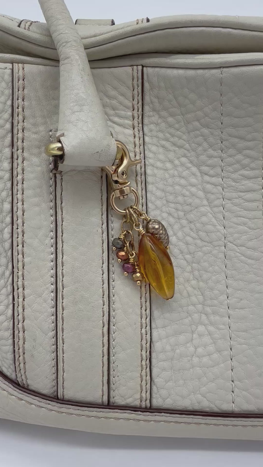 A video of a cream-colored leather bag featuring a decorative gold-tone clasp and charm with a large amber-colored faceted bead and small multicolor glass beads - made with love in Colorado by Ruby Acorn.