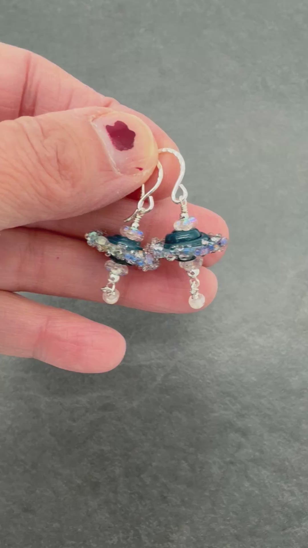 A video of a hand holding a pair of handmade dangle earrings with silver fishhooks, featuring dark blue lampwork glass beads, iridescent clear beads, and dangling clear faceted beads.
