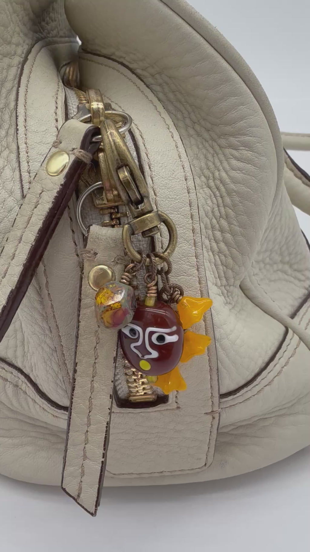 Video close-up of a cream-colored textured leather handbag featuring a brass-tone zipper pull adorned with decorative glass charms, including a brown face pendant and orange-yellow beads - made with love in Colorado by Ruby Acorn.