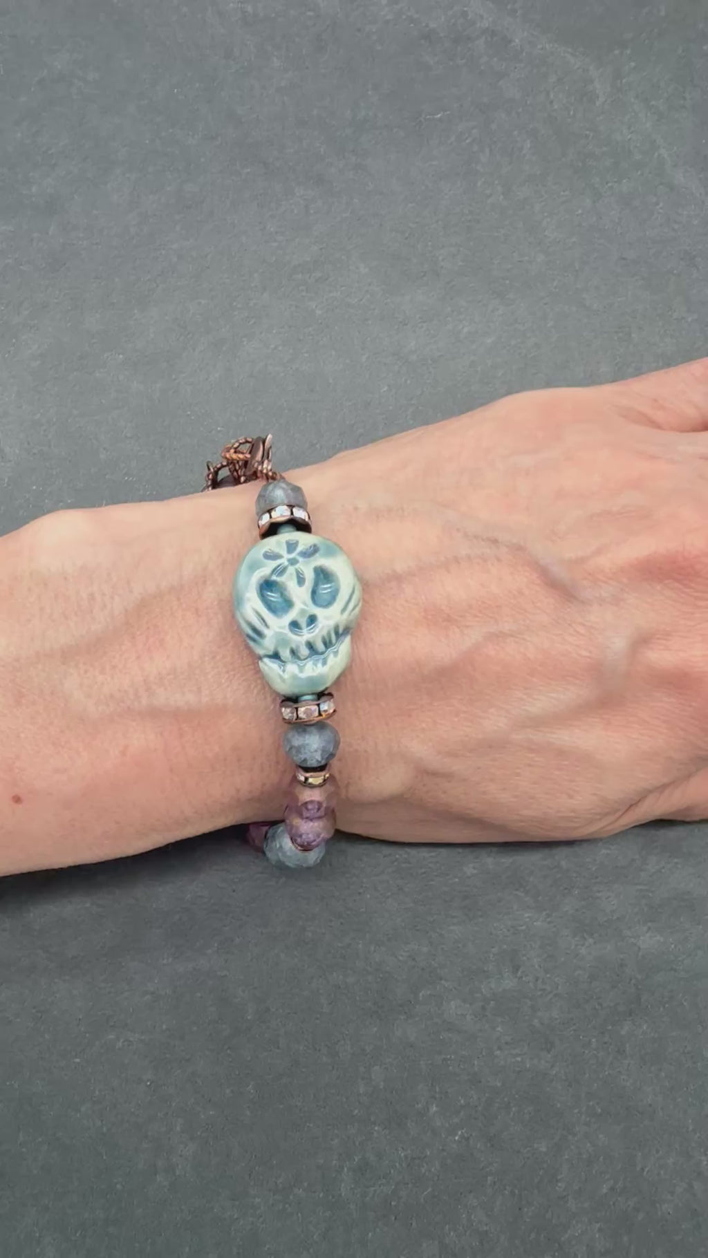 Video of a beaded bracelet with a blue Day of the Dead sugar skull and dark beads worn on a person's wrist, shown against a dark gray textured background - made with love in Colorado by Ruby Acorn.