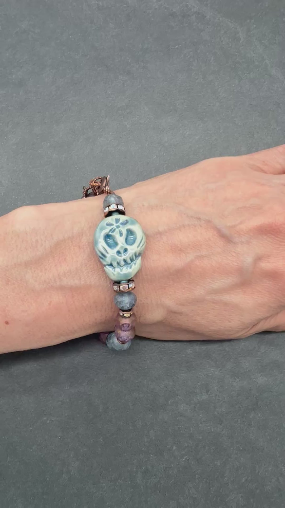 Video of a beaded bracelet with a blue Day of the Dead sugar skull and dark beads worn on a person's wrist, shown against a dark gray textured background - made with love in Colorado by Ruby Acorn.
