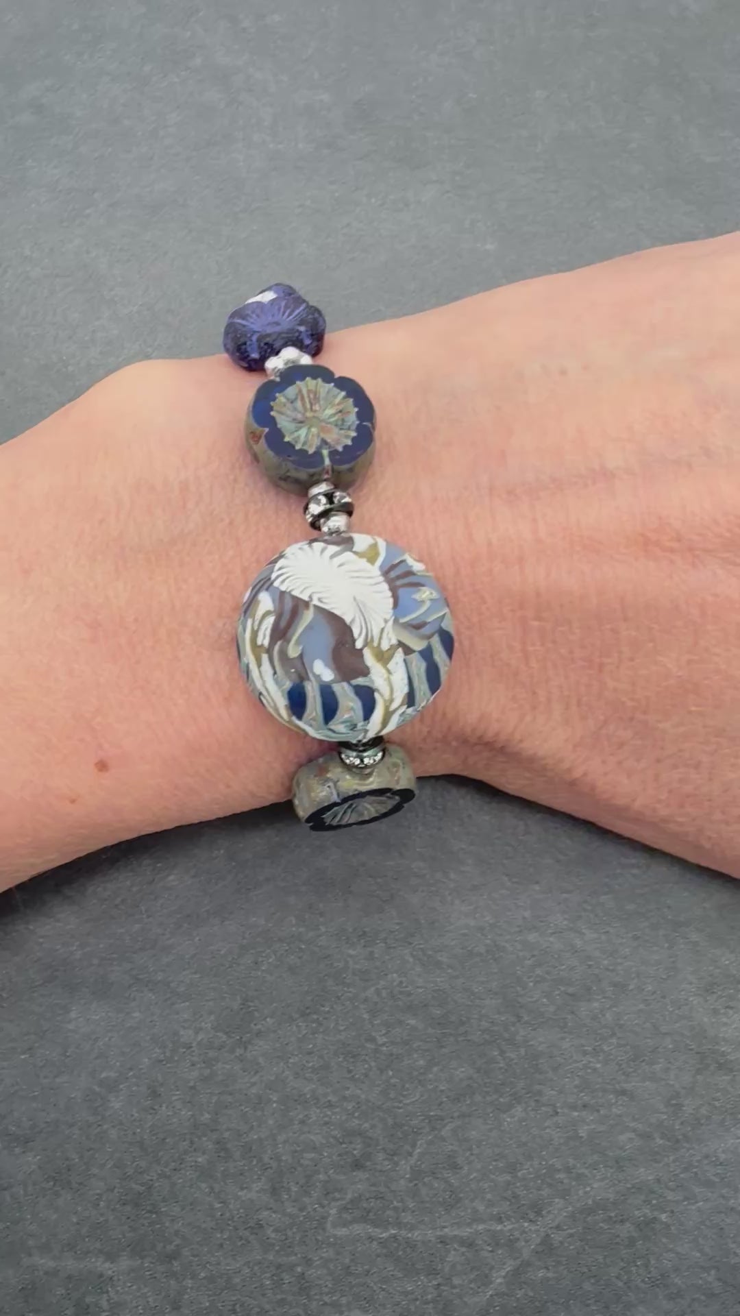 A video of a beaded bracelet with dark blue, and silver-toned beads, including a large polymer clay bead, shown on a person's wrist against a dark gray textured background.
